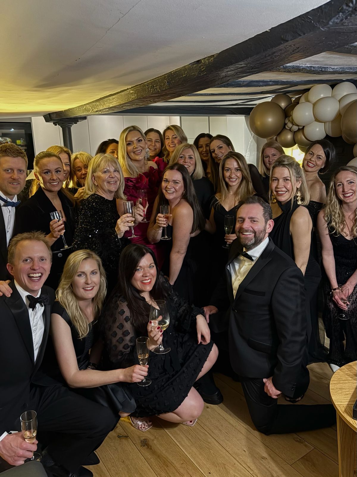 A large group in black tie raising glasses under the beams, beside a gold balloon arch